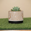A cylindrical ceramic pot with a light wood grain patterned body and a matte black base, sitting on a strip of green artificial grass.