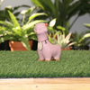 A small pink llama figurine with orange accents and a cream-colored heart-shaped tag, standing on artificial green grass against a plain white wall.