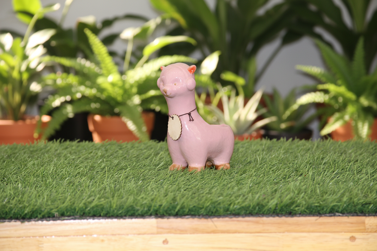 A small pink llama figurine with orange accents and a cream-colored heart-shaped tag, standing on artificial green grass against a plain white wall.