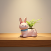 A small pink bunny figurine with a blue patterned collar sits on a strip of artificial green grass, placed on a wooden surface against a plain light gray wall.