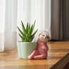 A small pink sloth figurine wearing a yellow hat sits beside a pale blue-green planter on artificial green grass, with a wooden surface below and a plain light gray wall behind.