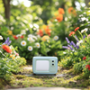 A miniature light blue toy television set with a blank white screen, placed on a strip of green artificial grass above a wooden surface against a plain off-white wall.