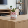 A small owl-shaped ceramic figurine with red and white patterns, sitting on a strip of green artificial grass above a wooden surface against a plain light gray background.