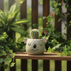 A small ceramic koala head planter in a pale sage green color, featuring a smiling face and pink inner ears, placed on a strip of green artificial grass above a wooden surface.
