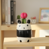 A small ceramic panda-shaped container or planter with a black and white body, standing on artificial green grass against a plain light wall.