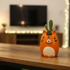 An orange and white ceramic cat-shaped container or planter, styled sitting on a strip of green artificial grass against a plain light gray wall.